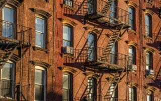 Close-up van de gevel van een klassiek rood stenen appartementencomplex in New York, met meerdere niveaus van gietijzeren brandtrappen die als externe vluchtwegen dienen.